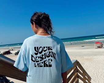 May include: A person wearing a light blue t-shirt with the text "YOUR DESIGN HERE" printed on the back. The person is standing on a wooden boardwalk overlooking a beach.