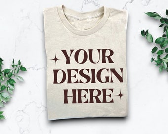 Puede incluir: Una camiseta beige doblada con un gráfico marrón que dice "YOUR DESIGN HERE" con una estrella a cada lado del texto.