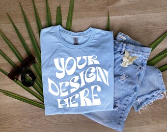 May include: A light blue t-shirt with the text "YOUR DESIGN HERE" in white, paired with light blue denim shorts. The image includes sunglasses, a scrunchie, and a decorative flower, all arranged on a wooden surface with green palm leaves.