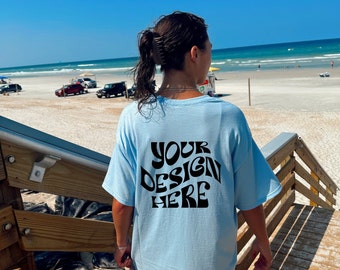 May include: A light blue t-shirt with the text "YOUR DESIGN HERE" printed in black on the back. The shirt is being worn by a person standing on a wooden staircase overlooking a beach.