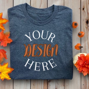 May include: A folded, dark gray t-shirt with the words "YOUR DESIGN HERE" in white and orange script. The shirt is surrounded by fall-themed decorations, including orange and yellow leaves and small pumpkins, on a wooden background.