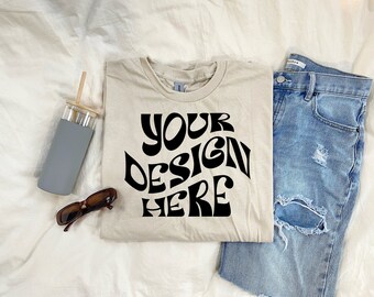 Puede incluir: Una camiseta beige con el texto "YOUR DESIGN HERE" impreso en negro en una fuente retro. La camiseta está colocada sobre una superficie blanca con unas gafas de sol, una botella de agua gris y unos vaqueros azules.