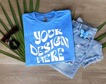 May include: A light blue t-shirt with the text "YOUR DESIGN HERE" in white,  folded on a wooden surface with a pair of blue denim shorts and a blue butterfly accessory.