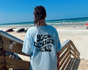 May include: A person wearing a light blue t-shirt with the text "YOUR DESIGN HERE" printed on the back. The person is standing on a wooden staircase overlooking a beach.
