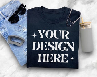 May include: A black t-shirt with the text "YOUR DESIGN HERE" in white letters. The shirt is folded and laid on a white marble surface with a pair of blue jeans, sunglasses, a hair clip, a necklace, and a gray water bottle with a bamboo lid.