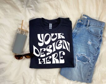 May include: A black t-shirt with the text "YOUR DESIGN HERE" in white,  psychedelic font. The t-shirt is folded and laid on a beige surface with a pair of sunglasses, a gray water bottle, and a pair of blue jeans.