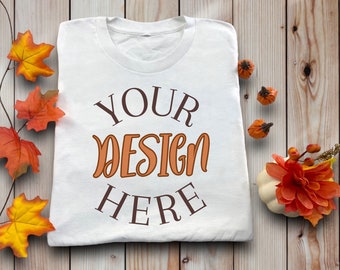 May include: White t-shirt with the text "YOUR DESIGN HERE" in brown and orange lettering. The shirt is on a wooden background with fall-themed decorations, including small pumpkins and orange and yellow leaves.