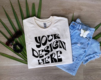 Puede incluir: Camiseta color crema con el texto "YOUR DESIGN HERE" en negro, combinada con shorts vaqueros azul claro y accesorios. Los shorts tienen detalles desgastados. También hay gafas de sol y una goma para el pelo.