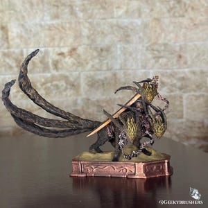 May include: A detailed figurine of a mythical creature with three heads, a long tail, and a sword. The sculpture is primarily brown and gold, with intricate detailing. The base is rectangular and bronze-colored. The overall theme is fantasy.