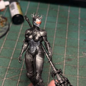 May include: A metallic dark gray humanoid figurine featuring a textured, armored suit, a crown-like headpiece, and a bright orange, faceted visor. The figure holds a long, chain-adorned weapon in one hand and stands in a rigid, upright posture.