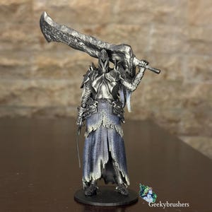 May include: A detailed figurine of a knight in silver armour, holding a large sword overhead. The knight wears a blue and grey robe with tattered edges. The base is black, and the background is a textured, light brown wall.