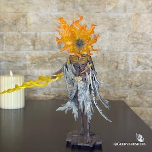 May include: A detailed figurine of a humanoid figure with a sun-like head and flowing orange and yellow flames. The figure is draped in tattered, gray fabric and holds a yellow, lightning-like object. The base is a wooden platform.