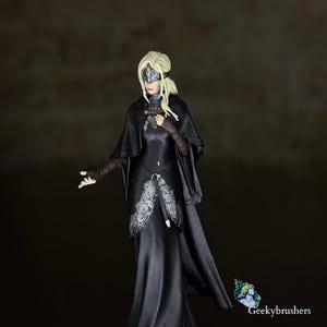 Puede incluir: Figura de una mujer con un vestido largo negro con detalles de encaje plateado. Lleva una capa negra, una venda en los ojos y tiene el pelo rubio. La figura está posando con un brazo extendido. El texto "Geekybrushers" está en la esquina inferior derecha.