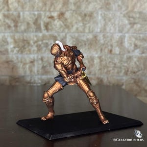 May include: A miniature figurine of a knight in bronze-colored armor, posed in a dynamic stance. The knight is holding a sword, with a white plume on the helmet. The figurine is mounted on a black base.