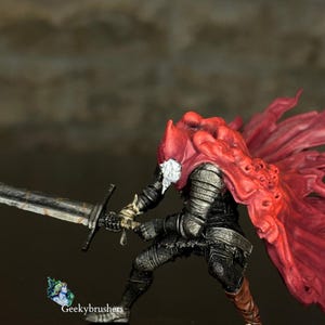 May include: A miniature figurine of a knight in dark armor, wielding a large sword. The knight wears a red hooded cloak with a white collar. The figurine is posed in a dynamic action stance, ready for battle.