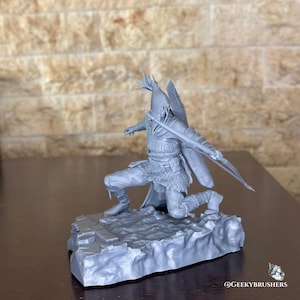 May include: A gray figurine of a kneeling archer with a bow and arrow, wearing detailed armor and a helmet with feather-like protrusions. The figure is posed on a rocky base, suggesting a fantasy or historical theme.