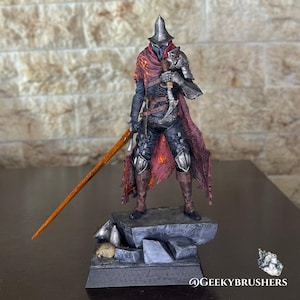 Puede incluir: Figura detallada de un guerrero con armadura oscura, capa roja y casco puntiagudo, empuñando una espada y una daga. La figura está sobre una base de piedra. La espada tiene una hoja naranja. Objeto de colección.