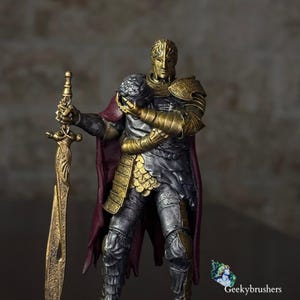 Op de afbeelding: Een gedetailleerd ridderbeeldje in versierd goud en zilverkleurig harnas, met een lang, ingewikkeld gouden zwaard in de ene hand en een helm in de andere. De ridder draagt een dieprode cape en heeft complexe metalen texturen en gravures.