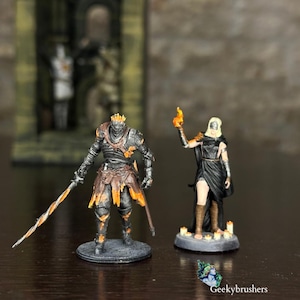 May include: Two detailed miniature figurines. One is a knight in black and orange armor, holding a sword. The other is a woman in a black dress, holding a flame. Both are on circular bases, with a dark brown background.