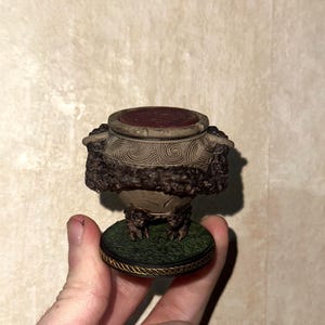 May include: A miniature decorative urn with a dark red top and intricate carvings. The urn has a textured, brown body and sits on a green base with a gold trim. The base has a grass-like texture.
