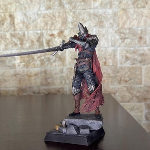 Könnte beinhalten: Detaillierte Figur eines Ritters in dunkler Rüstung, der ein langes Schwert hält. Der Ritter trägt einen roten Umhang und steht auf einem Steinsockel mit Totenköpfen. Die Figur ist mit aufwendigen Details bemalt und zeigt ein mittelalterliches Fantasy-Thema.