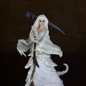Pode incluir: Uma estatueta branca de uma figura feminina com longos cabelos brancos e um vestido esvoaçante, segurando uma foice. A figura tem uma tez pálida e olhos escuros. A foice é cinza e o bastão é marrom.