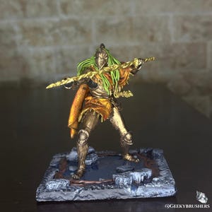 Może przedstawiać: Miniaturowa figurka wojownika w złotej zbroi, trzymającego złotą broń. Wojownik ma zielone włosy i pomarańczową pelerynę. Figurka stoi na kamiennej podstawie.