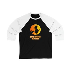 Pode incluir: Camiseta de beisebol preta e branca com um gráfico branco e laranja de uma bruxa voando em uma vassoura na frente de uma lua cheia. O texto "STAY SPOOKY, WITCHES" é impresso em laranja abaixo do gráfico.