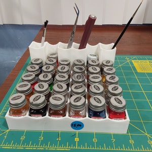 May include: A white 3D-printed organizer holding small glass bottles of paint with silver lids. The organizer also holds paintbrushes, tweezers, and a stylus. The bottles are arranged on a green cutting mat.