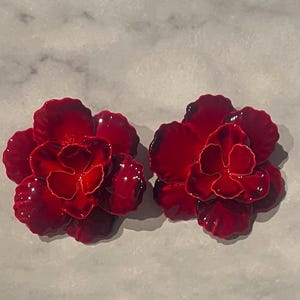 May include: Two vibrant red flower-shaped earrings. Each earring features multiple petals and a glossy finish, with a darker red center. The earrings are designed to resemble a blooming flower, adding a touch of nature-inspired elegance.