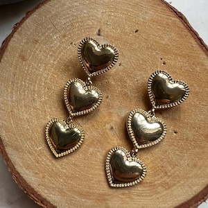 May include: A pair of gold-tone dangle earrings with three heart-shaped charms. Each heart has a textured surface and a beaded edge.