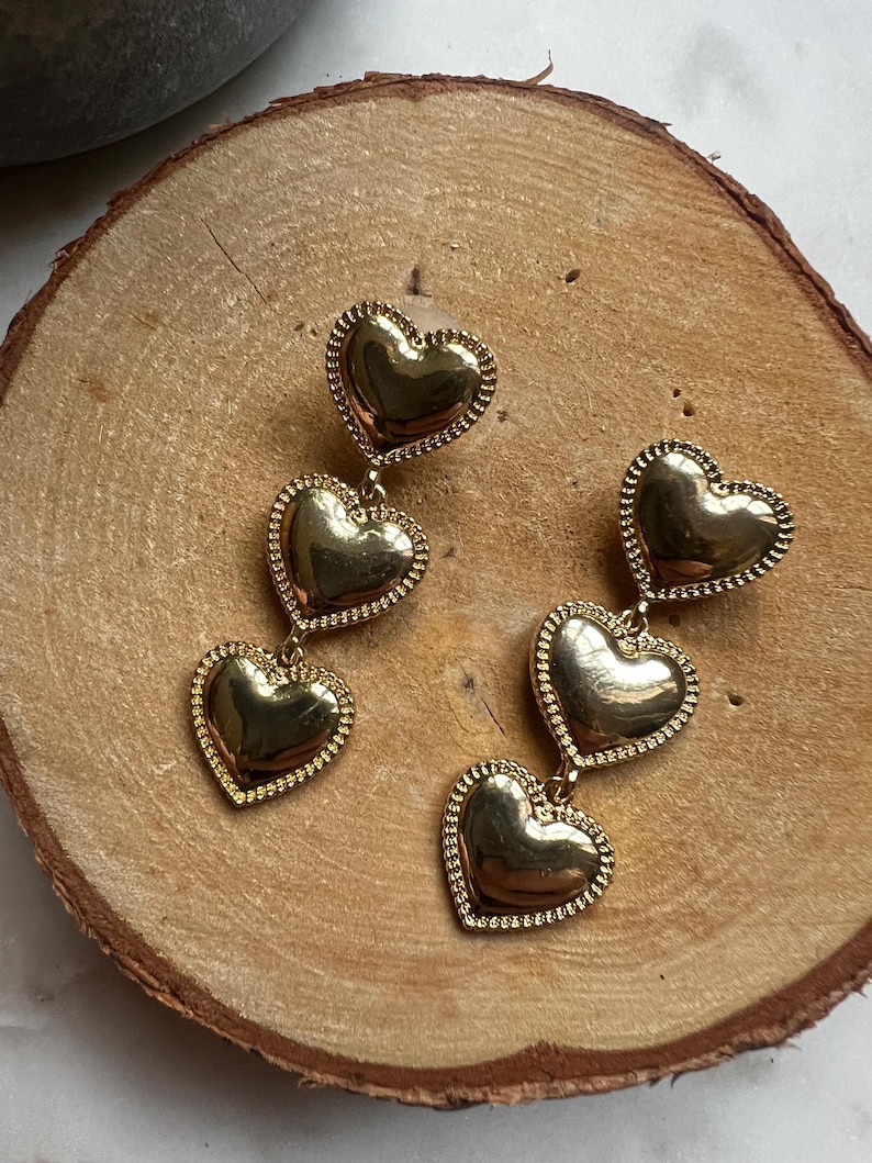May include: A pair of gold-tone dangle earrings with three heart-shaped charms. Each heart has a textured surface and a beaded edge.