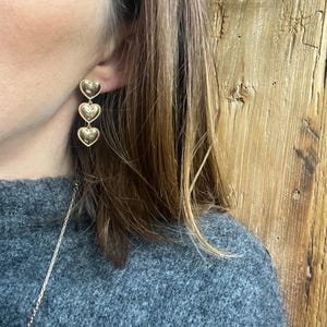 May include: Gold heart shaped dangle earrings with three hearts hanging from each other. The earrings are worn by a person with long brown hair.