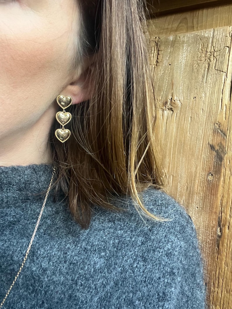 May include: Gold heart shaped dangle earrings with three hearts hanging from each other. The earrings are worn by a person with long brown hair.