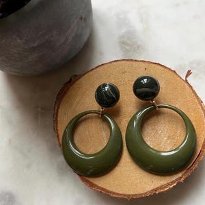 Puede incluir: Pendientes de aro verde oliva con un perno oscuro similar al mármol. Los pendientes se exhiben sobre una rodaja de madera, mostrando su acabado brillante y su diseño sencillo. Los pendientes tienen un poste de color dorado.