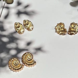 May include: Three pairs of gold-colored stud earrings. The earrings are in the shape of seashells. One pair is oval, one pair is a spiral, and the third pair is a rose shape. The earrings are on a white surface.