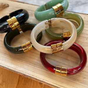 May include: A selection of colourful, chunky resin bangles. The bracelets are in shades of black, green, white, red, and olive green. Each bangle has a gold-coloured clasp. The bangles are displayed on a wooden tray.