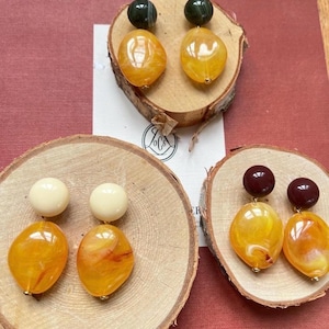 May include: Three pairs of handmade earrings. Each pair features a different combination of colors and shapes. One pair has dark green and yellow-orange stones, another has cream and yellow-orange stones, and the last has dark red and yellow-orange stones.