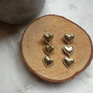 May include: A pair of gold-tone earrings with three heart-shaped charms hanging from each earring.