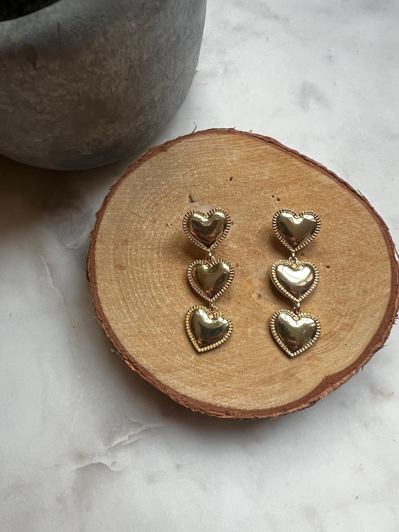 May include: A pair of gold-tone earrings with three heart-shaped charms hanging from each earring.