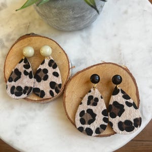 Puede incluir: Dos pares de pendientes colgantes de estampado de leopardo. Un par tiene manchas beige y negras con una cuenta beige claro en la parte superior. El otro par tiene manchas beige y negras con una cuenta negra en la parte superior.