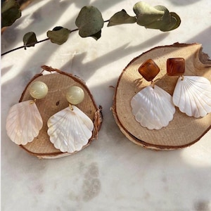 Op de afbeelding: Twee paar oorbellen met schelpmotieven. Eén paar heeft een lichtgroene bol en een roze schelp. Het andere paar heeft een bruin vierkant en een witte schelp. Beide paren liggen op houten schijfjes.