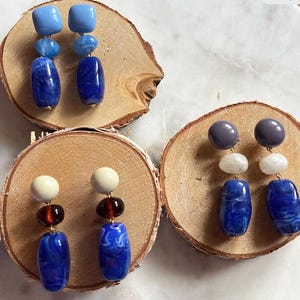Op de afbeelding: Een collectie handgemaakte oorbellen met verschillende kleuren en ontwerpen. De oorbellen hebben blauwe, witte en bruine kralen, met goudkleurige hardware. De oorbellen worden gepresenteerd op kleine houten schijfjes.
