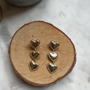 May include: Gold tone dangle earrings with three heart-shaped charms. The hearts are layered and have a textured surface.
