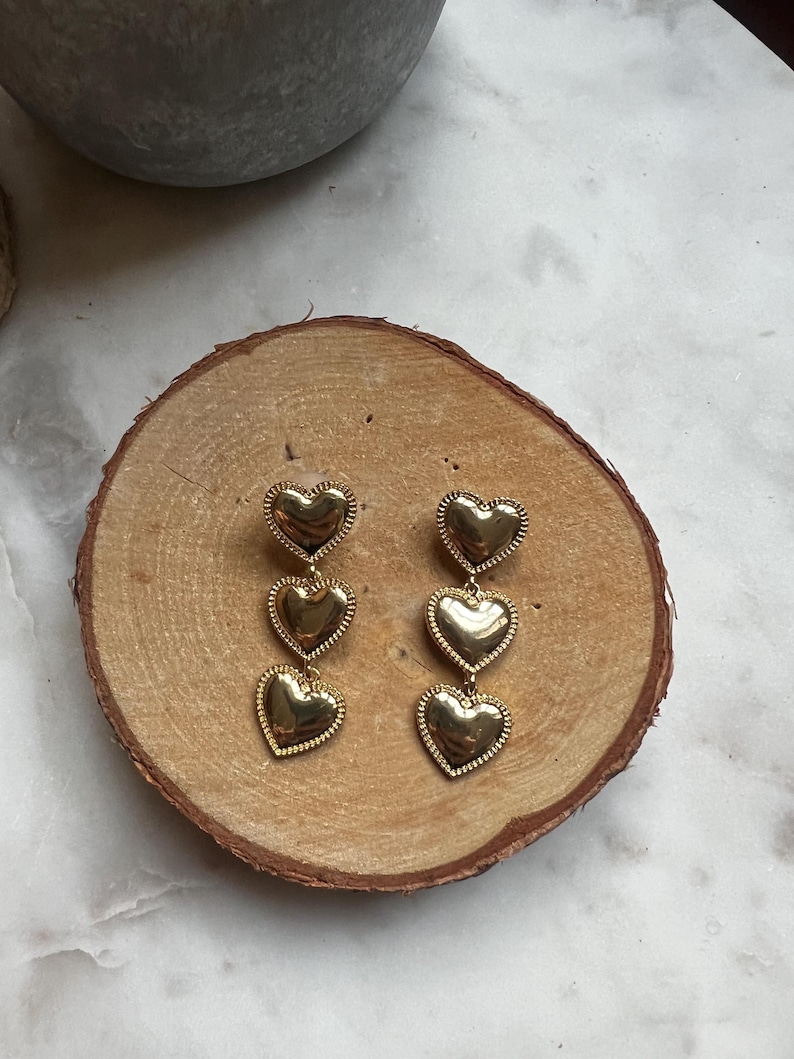 May include: Gold tone dangle earrings with three heart-shaped charms. The hearts are layered and have a textured surface.