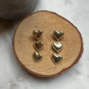May include: Gold tone dangle earrings with three heart shaped charms. The hearts are layered and have a textured surface.