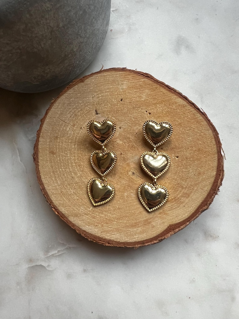May include: Gold tone dangle earrings with three heart shaped charms. The hearts are layered and have a textured surface.