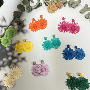 Peut inclure: Diverses boucles d'oreilles en forme de marguerites, aux couleurs vives : orange, rose, bleu, vert, jaune et lavande. Les boucles d'oreilles sont disposées sur une surface blanche, avec des brins d'eucalyptus. Chaque paire a un petit clou rond.