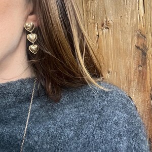 May include: Gold heart-shaped earrings with three hearts hanging in a row. The earrings are worn by a person with long brown hair.