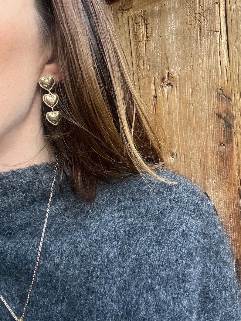 May include: Gold heart-shaped earrings with three hearts hanging in a row. The earrings are worn by a person with long brown hair.